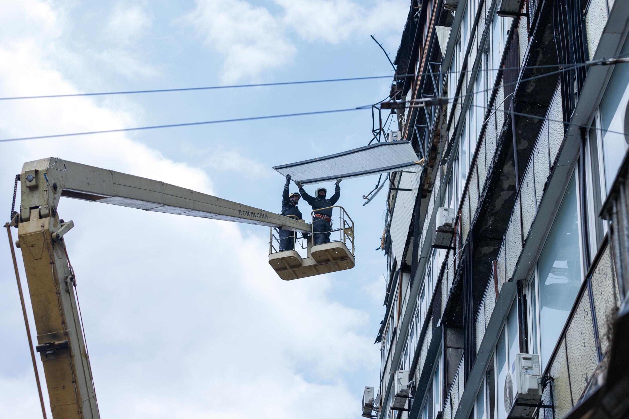 Двоє працівників у захисному одязі стоять у підйомному кошику спецтехніки біля фасаду багатоповерхового будинку. Вони демонтують пошкоджену частину облицювання або даху. На будівлі помітні ознаки руйнувань.