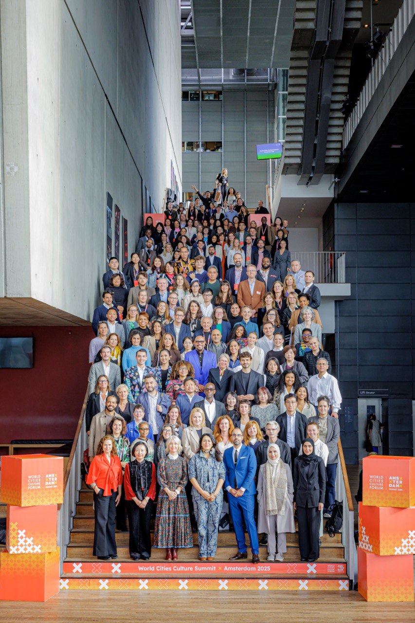 Групове фото учасників Всесвітнього саміту культури міст World Cities Culture Summit 2025 в Амстердамі. Велика група людей стоїть на сходах у сучасній будівлі з бетонними стінами та скляними конструкціями. На передньому плані — банери з написом World Cities Culture Forum. Amsterdam 2025. Учасники різного віку та статі позують для спільного фото, більшість усміхаються.