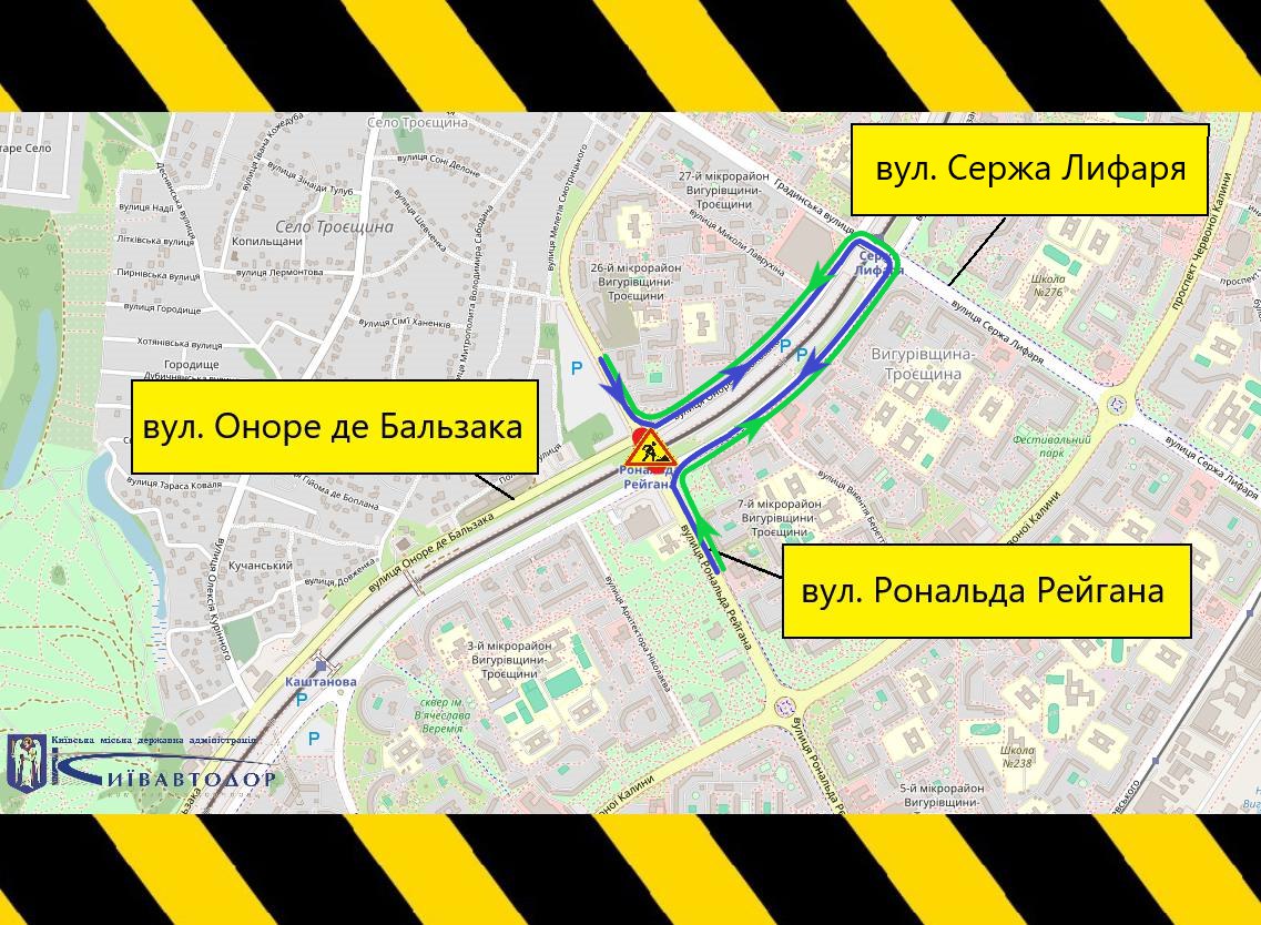 Схема об’їзду в місті Київ (район Троєщина). Перекриту ділянку позначено на перехресті вулиць вул. Оноре де Бальзака та вул. Рональда Рейгана. Об’їзд організовано по кільцю через вул. Сержа Лифаря: рух із боку вулиці Оноре де Бальзака спрямовується праворуч на вулицю Рональда Рейгана; далі транспорт рухається вгору по вулиці Сержа Лифаря; після цього повертає ліворуч і повертається на вулицю Оноре де Бальзака, минаючи закриту ділянку. Напрямки руху позначені зеленими та синіми стрілками. У центрі схеми — знак дорожніх робіт. По краях зображення — чорно-жовта попереджувальна рамка. Внизу ліворуч розміщено логотип «Київавтодор».