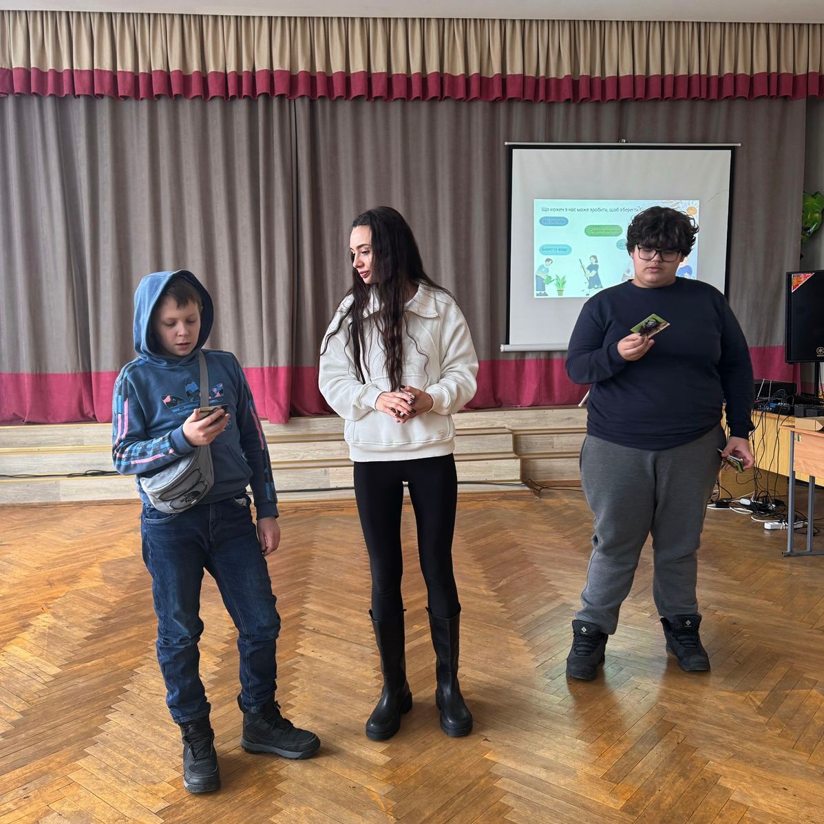 На світлині троє людей - двоє підлітків та молода жінка - стоять у залі з паркетною підлогою на фоні сцени з коричневими завісами. Зліва хлопець у синьому худі з капюшоном дивиться у свій телефон, а праворуч інший хлопець у темному светрі тримає в руках невелику картку. Посередині стоїть жінка у білій кофті та чорних штанах, яка спостерігає за діями підлітків. Праворуч на задньому плані видно проєкційний екран з презентацією, де зображені ілюстрації та текст. 