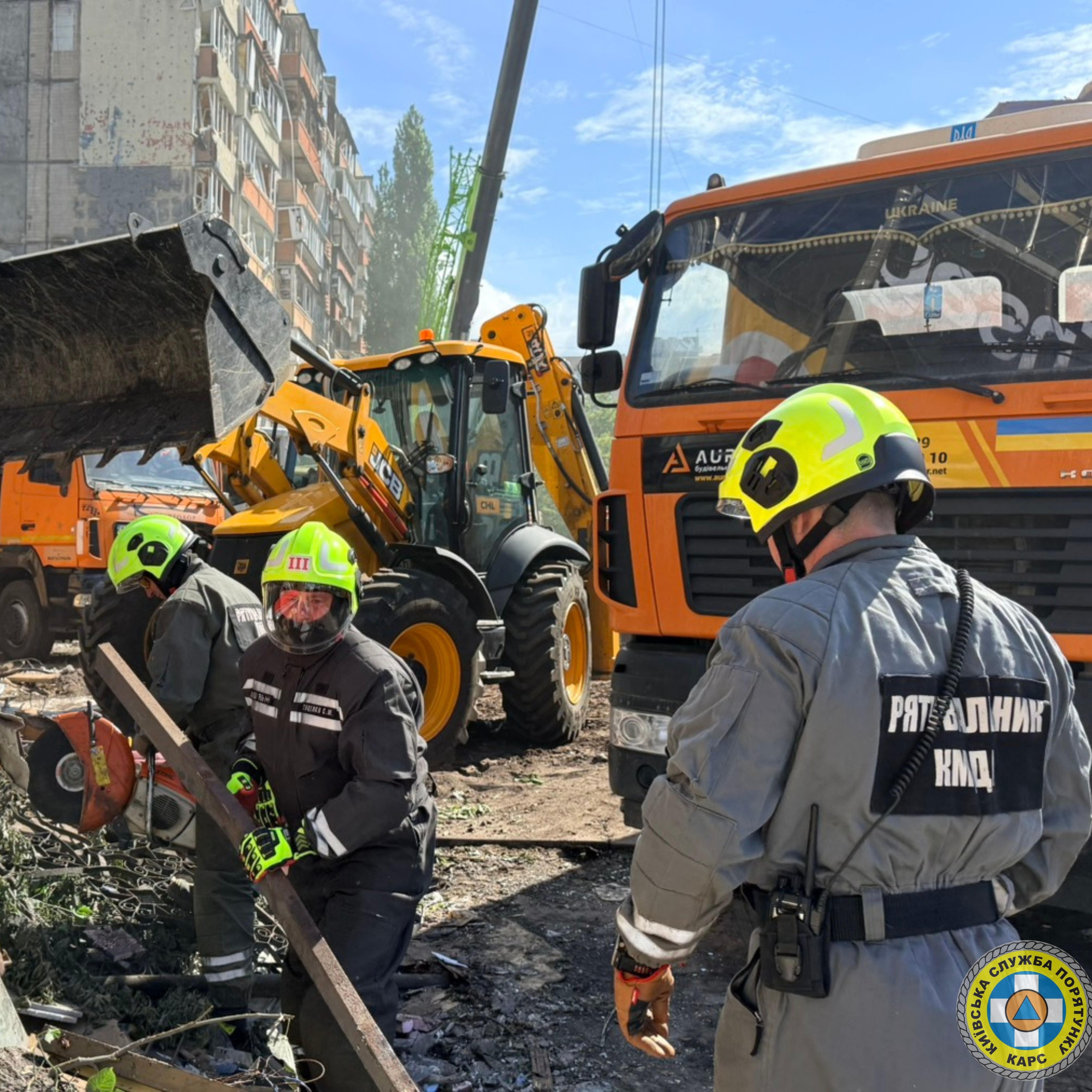 На світлині зображено роботу рятувальників «Київської служби порятунку» на місці руйнувань у місті. На передньому плані троє чоловіків у сірій формі та яскраво-жовтих захисних шоломах розбирають завали вручну. Один із них тримає важку металеву балку, а на спині іншого видно напис «РЯТУВАЛЬНИК КАРС». Безпосередньо за ними стоїть важка спецтехніка: велика помаранчева вантажівка та жовтий екскаватор із піднятим ковшем. На дальньому плані ліворуч височіє багатоповерховий житловий будинок із помітними слідами пошкоджень від обстрілу. 