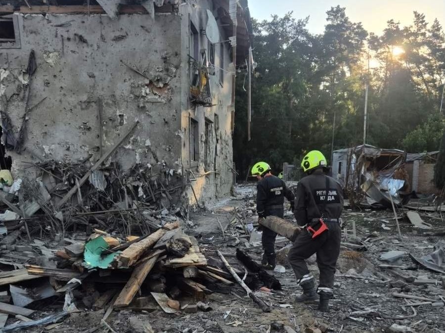Рятувальники працюють серед завалів біля багатоповерхового житлового будинку, пошкодженого внаслідок обстрілу. Фасад будівлі посічений уламками, частина конструкцій зруйнована. На передньому плані двоє рятувальників у захисному спорядженні розбирають уламки деревини та будівельного сміття. На задньому плані видно пошкоджені споруди та дерева.