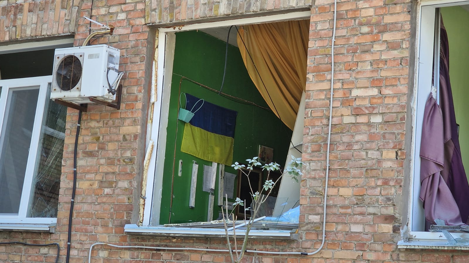 На фото — фасад цегляної будівлі з вибитим вікном. Усередині приміщення видно зелено пофарбовані стіни, український прапор на стіні та пошкоджені меблі. На підвіконні лежать уламки скла й рами, частина віконної конструкції зруйнована. Поруч із вікном на фасаді встановлений зовнішній блок кондиціонера, а в сусідніх вікнах також помітні пошкодження.