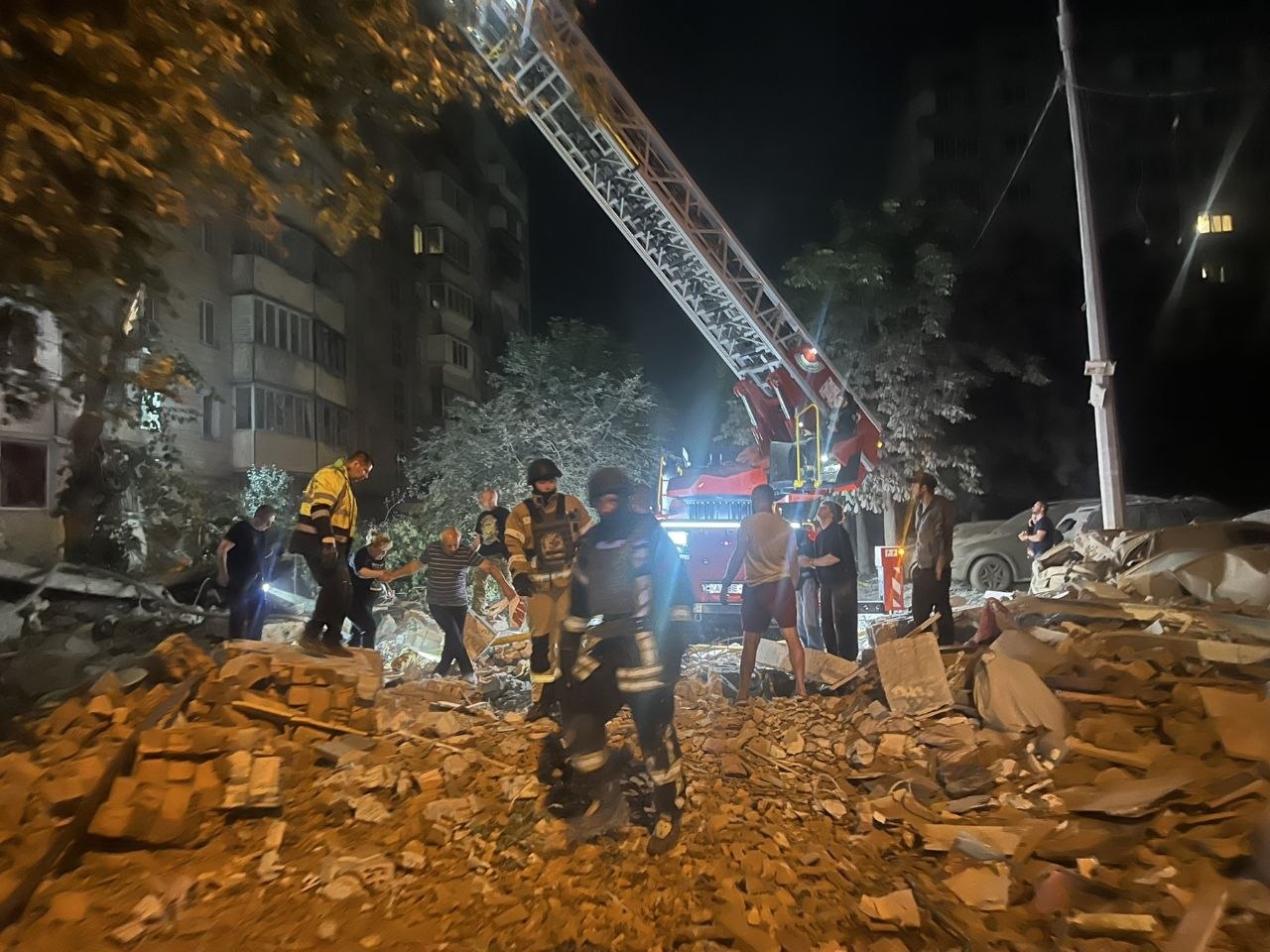У нічний час рятувальники та комунальні служби працюють на місці руйнувань біля багатоповерхового житлового будинку. На передньому плані — великі завали з уламків бетону та цегли, серед яких пересуваються рятувальники в захисному спорядженні й місцеві жителі. Позаду працює пожежний автопідіймач із висунутою стрілою, територію освітлює спецтехніка. Тривають аварійно-рятувальні роботи після ворожої атаки.